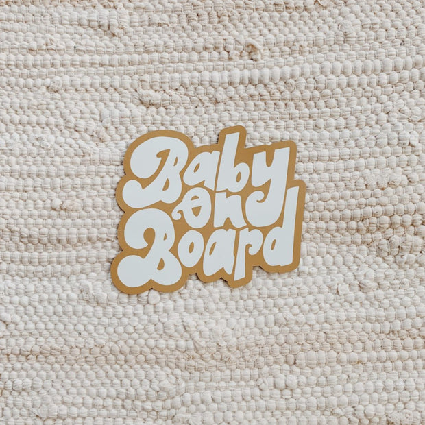 A car magnet reads "Baby on Board" in white and ochre text, it sits on top of a cream woven rug