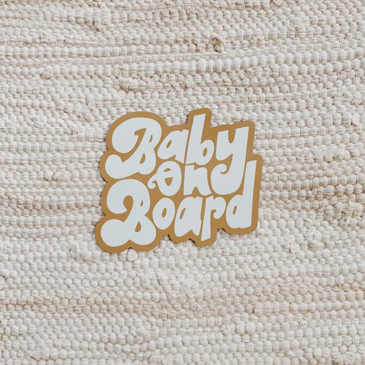 A car magnet reads "Baby on Board" in white and ochre text, it sits on top of a cream woven rug